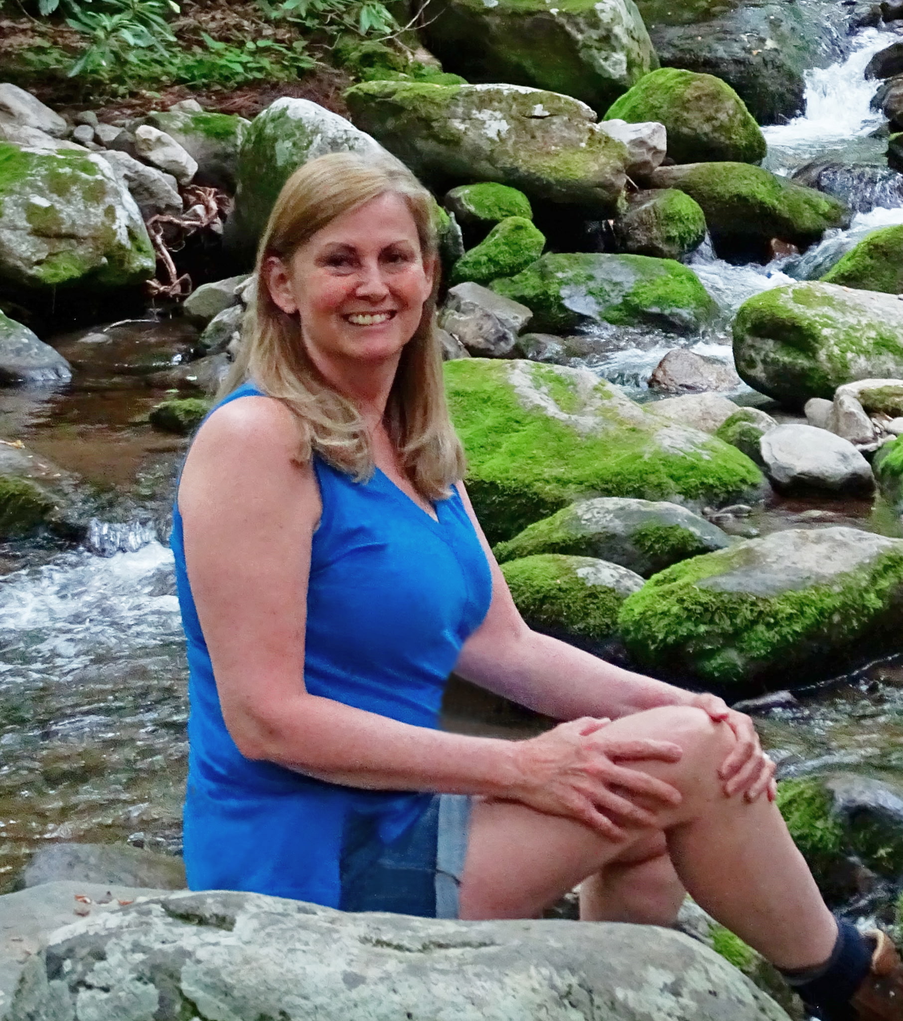 A picture of Anne Tully sitting beside moss covered rocks and a flowing creek